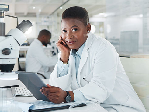 woman in white lab coat sitting at a table looking at a tablet